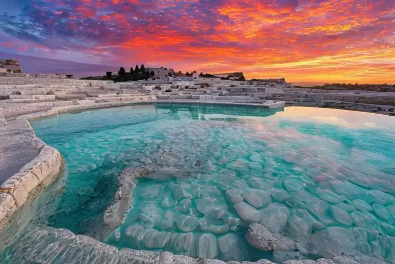 A panoramic view of the white travertine terraces of Pamukkale, Turkey, filled with clear turquoise thermal waters, with a dramatic, vibrant sunset painting the sky in fiery orange, pink, and purple hues, and the ancient ruins of Hierapolis visible in the distance on the hill.
