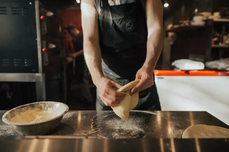 Chef pizzaiolo italiano artigiano che prepara una pizza napoletana fresca in una cucina professionale con forno a legna, farina in aria, movimento dinamico, alta qualità