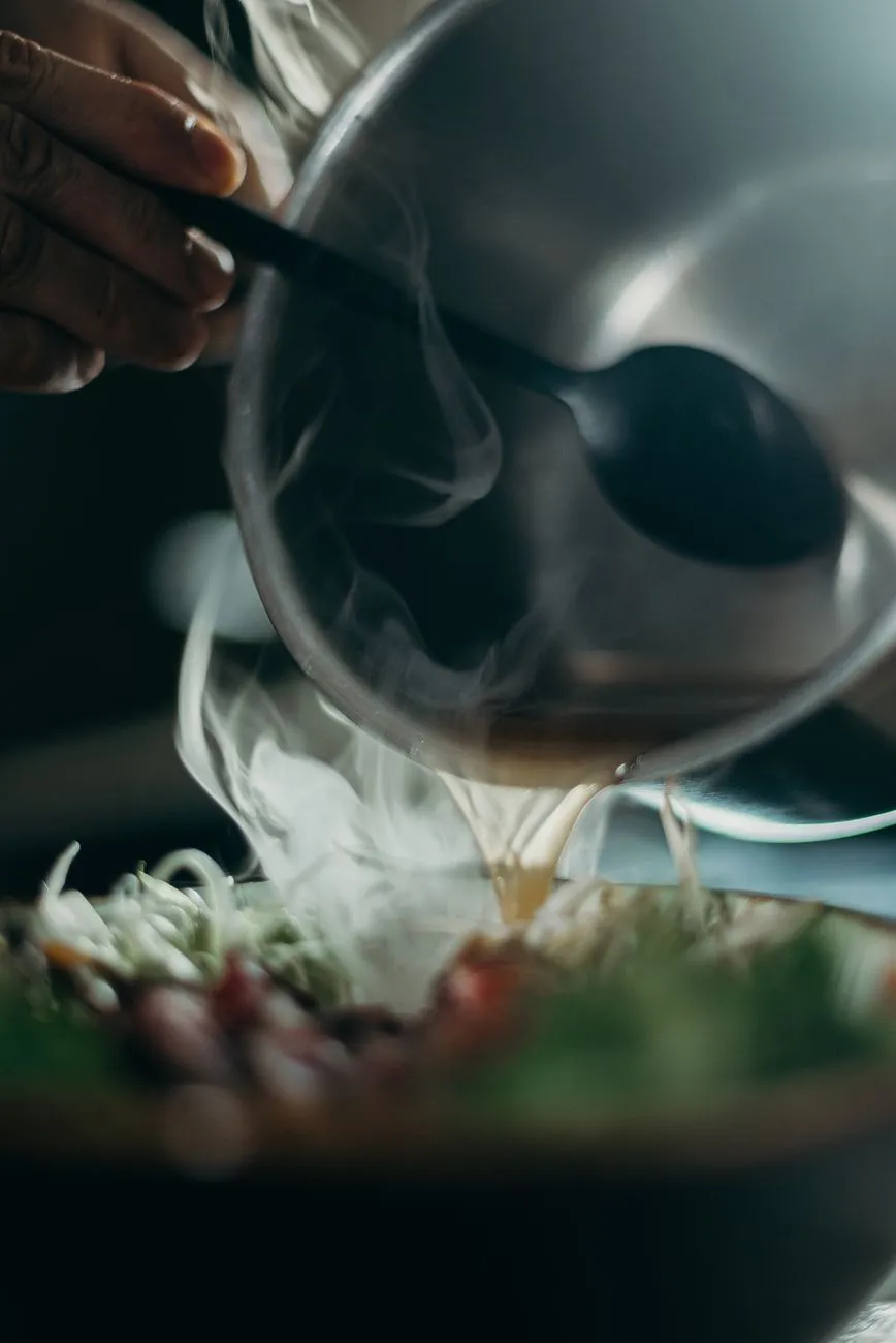 A chef ladling hot ramen broth into a bowl of noodles