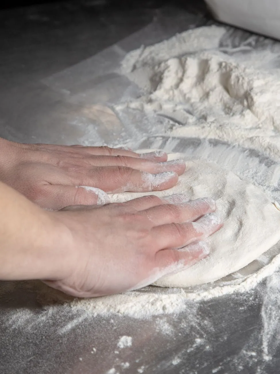 A baker's hands shaping a sourdough boule on a floured surface, with a bowl of starter nearby