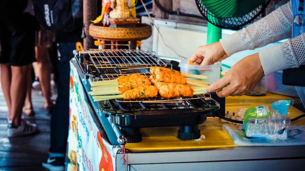 A bustling Bangkok street food stall with a vendor grilling satay skewers over hot coals