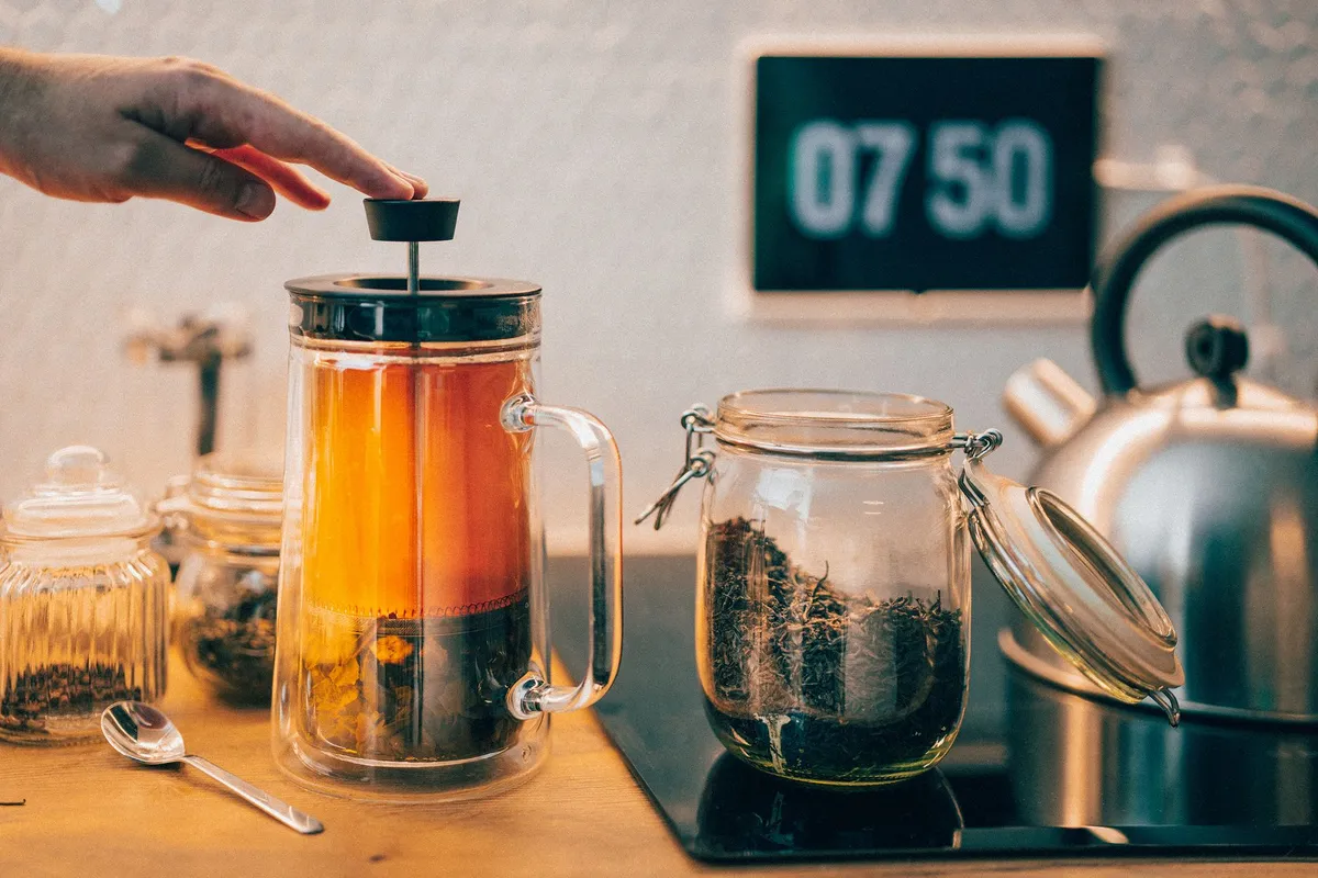 A glass jar filled with golden kombucha tea, topped with a thick SCOBY, surrounded by scattered tea leaves and lemon slices